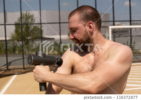 Shirtless man massaging shoulder with massage percussion device after workout on sports ground. Summer workout and training. Applying therapeutic percussive massager gun. Shirtless man massaging shoulder with massage percussion device after workout on sports ground. Summer workout and training. Applying therapeutic percussive massager gun. 116372877
