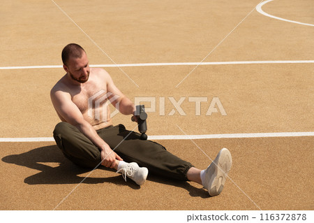 Shirtless man massaging leg with massage percussion device after workout on sports ground. Summer workout and training. Applying therapeutic percussive massager gun. 116372878