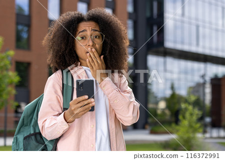 Young student with curly hair and glasses holding smartphone with shocked expression outdoor. Brick and glass building in background. Concept of surprise, technology, urban lifestyle. 116372991