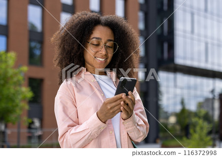 Smiling woman using smartphone outdoors in urban environment. Casual attire, relaxed atmosphere, focused on screen. Background of modern glass building. Smiling woman using smartphone outdoors in urban environment. Casual attire, relaxed atmosphere, focused on screen. Background of modern glass building. 116372998