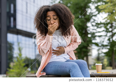 Young woman experiencing stomach pain and holding her hand over mouth. She is sitting outside in casual attire, indicating discomfort and possible nausea. 116373012