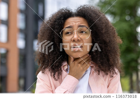 Young woman feeling discomfort due to sore throat standing outdoors. Person expressing pain and illness against blurred urban background. Health and medical concept Young woman feeling discomfort due to sore throat standing outdoors. Person expressing pain and illness against blurred urban background. Health and medical concept 116373018