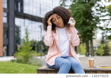 Young woman with curly hair and eyeglasses holding tissues while having headache. Sitting outside modern building, looking stressed. Concept of illness, stress, and urban lifestyle. 116373020