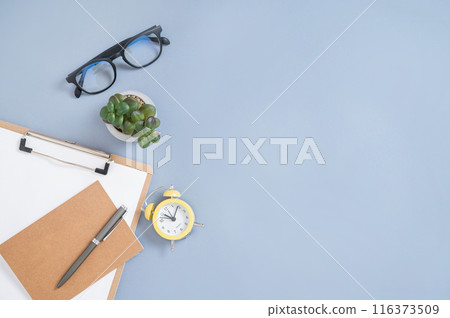 Flat lay craft notepad, pen, yellow alarm clock and eyeglass on a blue background.  116373509