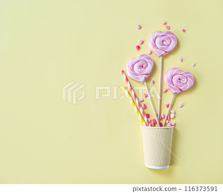 Pink lollipops in the shape of hearts at  paper cup with color sugar on a yellow background.  116373591