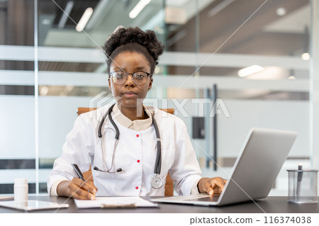 Confident female doctor working at desk in modern clinic. Doctor is using laptop and writing notes, demonstrating professionalism and dedication to medical practice. Confident female doctor working at desk in modern clinic. Doctor is using laptop and writing notes, demonstrating professionalism and dedication to medical practice. 116374038