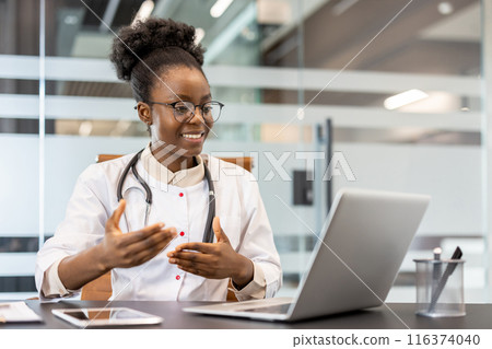 Professional doctor sitting at desk conducting video call consultation in modern office. Stethoscope around neck, gestures to communicate effectively. Medical professional using technology 116374040