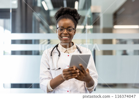 Confident doctor in white coat with stethoscope uses digital tablet in modern medical office. Medical professional looking at camera with smile, showcasing technology use in healthcare. 116374047