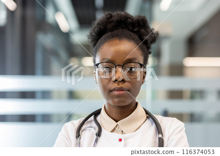 Female doctor wearing glasses and stethoscope standing confidently in modern medical office. Professional healthcare provider posing indoors, reflecting strength and competence 116374053