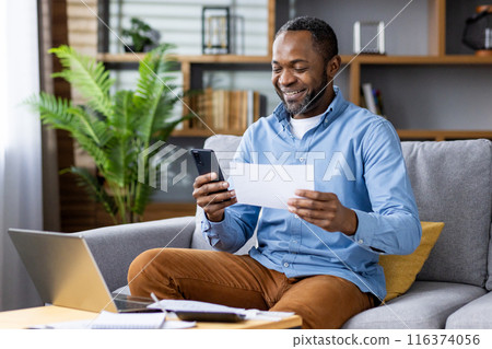 Smiling man working remotely from home, using smartphone and reviewing document while sitting on sofa with laptop and notepad. Concept of remote work home office, happiness, productivity, technology. 116374056
