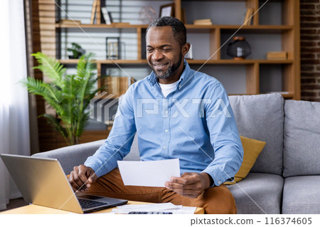 Smiling man enjoying remote work from home office while looking at financial documents. Entrepreneur managing business finances using laptop at home in cozy living room setting 116374605