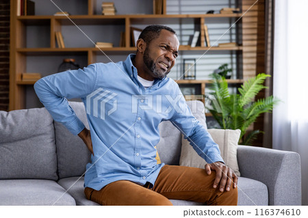 Middle-aged man experiencing back pain while sitting on comfortable couch in modern living room. Man grimacing in discomfort, holding back, in need of relief. Cozy, well-lit home interior setting. 116374610