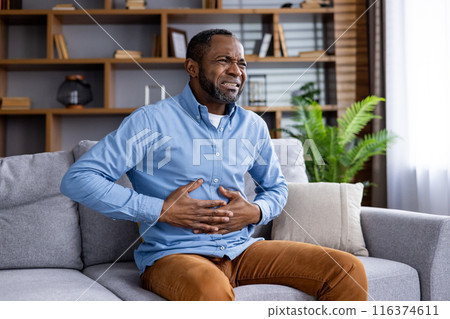 Man experiencing abdominal pain while seated on sofa in living room. Man clutching stomach due to feeling of discomfort. Concept of health, pain, and discomfort. 116374611