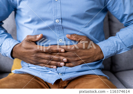 Close-up of man's hands clutching stomach indicating abdominal pain and discomfort. Concept of digestive issues, stomach ache, and health problems. Man in blue shirt and brown pants sitting on couch. Close-up of man's hands clutching stomach indicating abdominal pain and discomfort. Concept of digestive issues, stomach ache, and health problems. Man in blue shirt and brown pants sitting on couch. 116374614