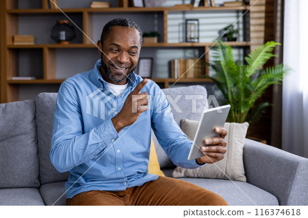 Happy man sitting on sofa at home having enjoyable video call using digital tablet. Smiling and relaxed man connecting with family or friends in comfortable living room setting. 116374618