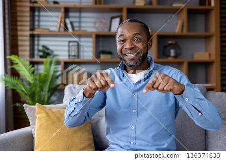Portrait of smiling man pointing at camera during video call while sitting in cozy living room. Joyful expressions, casual setting, indoor ambiance perfect for remote communication online conference. Portrait of smiling man pointing at camera during video call while sitting in cozy living room. Joyful expressions, casual setting, indoor ambiance perfect for remote communication online conference. 116374633