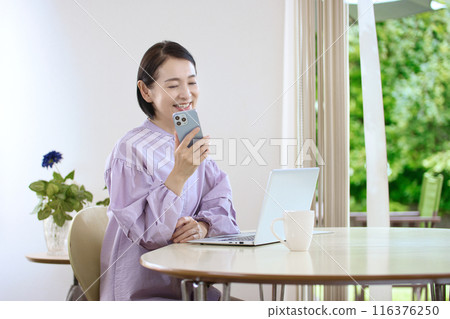A middle-aged woman looking at a computer while using her smartphone in the living room 116376250