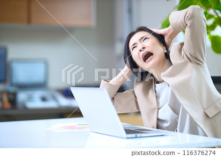 A middle-aged woman with a sour face holding her head in the office 116376274