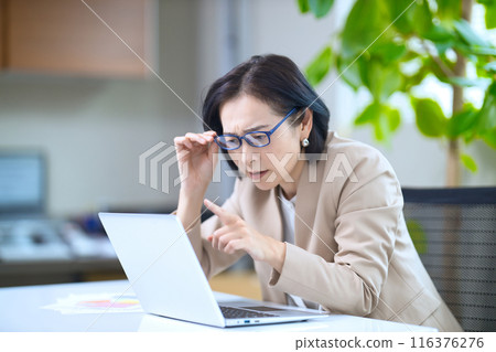 A middle-aged woman wearing reading glasses looking at a computer in the office A middle-aged woman wearing reading glasses looking at a computer in the office 116376276