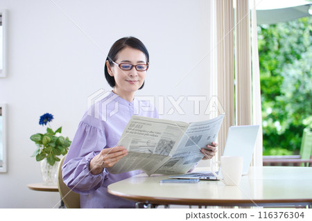 A middle-aged woman wearing reading glasses and reading an English newspaper 116376304