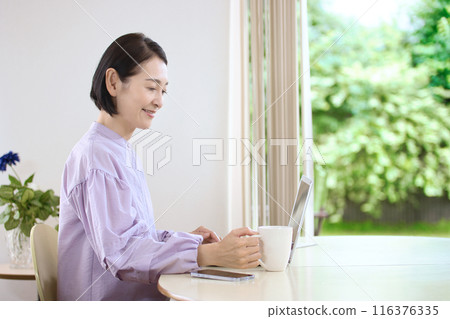A middle-aged woman drinking coffee and looking at a computer in the living room 116376335