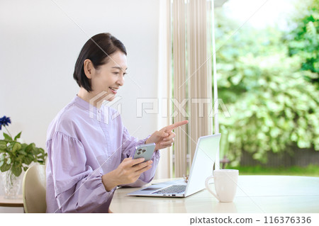 A middle-aged woman looking at a computer while using her smartphone in the living room A middle-aged woman looking at a computer while using her smartphone in the living room 116376336