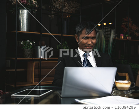 Smiling Businessman in suit working on laptop at dark, modern office desk with tablet and cup of coffee Smiling Businessman in suit working on laptop at dark, modern office desk with tablet and cup of coffee 116384299