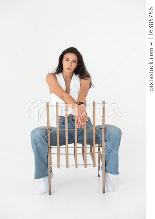 Happy beautiful girl posing with a chair in a new hot look. Wearing blue jeans and white waistcoat 116385756