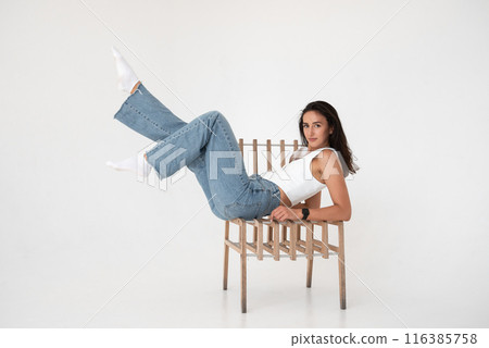 Happy beautiful girl posing with a chair in a new hot look. Wearing blue jeans and white waistcoat Happy beautiful girl posing with a chair in a new hot look. Wearing blue jeans and white waistcoat 116385758