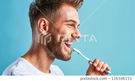 Young man in braces brushes his teeth, blue background copy space. Orthodontic device care concept, dental health, oral hygiene. 116386153