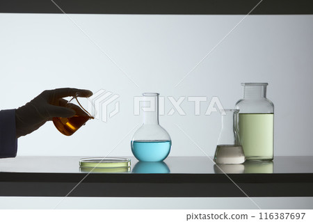 A beaker of yellow fluid held by a scientist hand, prepare to pour into a petri dish, which displayed on the table next to a boiling flask, erlenmeyer flask and glass bottle. Copy space for text A beaker of yellow fluid held by a scientist hand, prepare to pour into a petri dish, which displayed on the table next to a boiling flask, erlenmeyer flask and glass bottle. Copy space for text 116387697