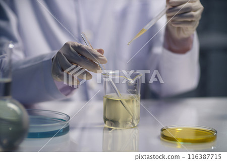 In the middle of the laboratory table displaying a glass beaker of light yellow fluid, the scientist is dripping yellow liquid from a dropper into a beaker below. The promotional photo, blank space In the middle of the laboratory table displaying a glass beaker of light yellow fluid, the scientist is dripping yellow liquid from a dropper into a beaker below. The promotional photo, blank space 116387715