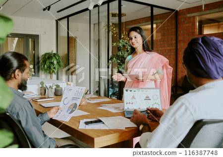 Young Indian startuper speaking to her colleagues and telling about financial statistics while male cowokers sitting at table and listening to her carefully 116387748