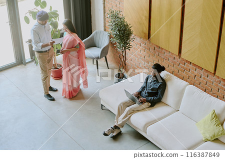 High angle shot of two cowokers chatting and discussing recent report while other sitting on sofa and programming 116387849