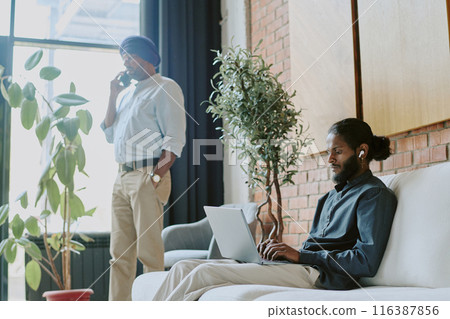 Low angle shot of two colleagues in comfortable coworking zone, one of them walking around room and speaking on phone while other one sitting on sofa and having online meeting 116387856