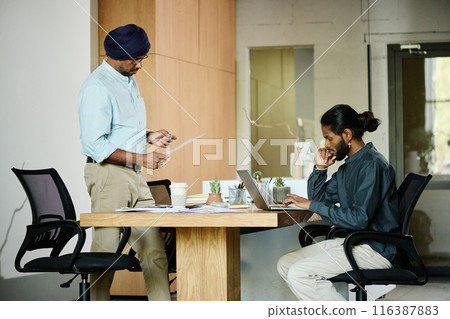 Two Indian coworkers having small conference, one of them reading report while other one searching information in Internet Two Indian coworkers having small conference, one of them reading report while other one searching information in Internet 116387883