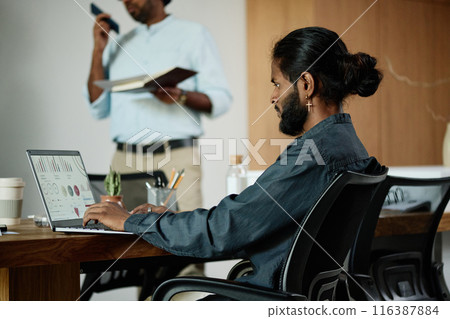 Young Indian IT specialist creating analytical presentation with colorful diagrams while his coworker speaking phone and holding his planner in background 116387884