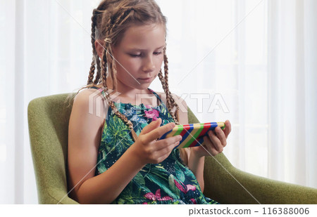 Teenage girl with lots of pigtails watches videos on her phone in room on chair. Technology and kids on school vacation. Teenage girl with lots of pigtails watches videos on her phone in room on chair. Technology and kids on school vacation. 116388006