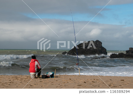 Fisherman on the sandy beach of the Atlantic Ocean in winter, man alone by the sea, beautiful cloudscape, dramatic landscape, gloomy seascape, big sea waves 116389294