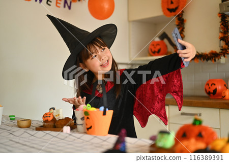 Smiling little girl in witch costumes taking selfies with decorated Halloween cupcakes in the kitchen Smiling little girl in witch costumes taking selfies with decorated Halloween cupcakes in the kitchen 116389391