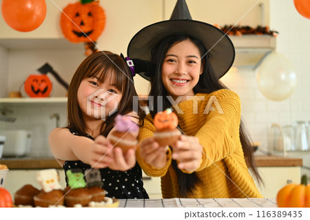Beautiful young mother and daughter showing Tasty Halloween cupcakes standing in decorated in the kitchen 116389435