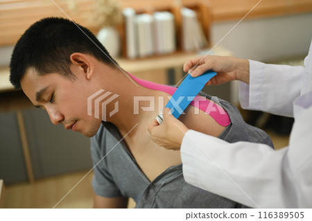 Physical therapist placing pink kinesio tape on man patient shoulder and neck at the clinic office. Alternative medicine concept. Physical therapist placing pink kinesio tape on man patient shoulder and neck at the clinic office. Alternative medicine concept. 116389505