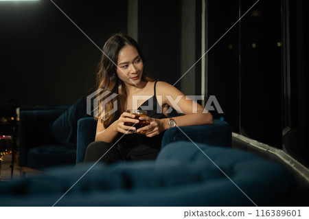 Elegant young woman in evening dress holding a drink sitting alone in a dimly lit lounge. Nightlife and luxury lifestyle concept 116389601