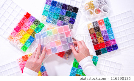 Flat lay. Woman hands gracefully poised over a collection of beads, sorted by color in transparent organizers. The array of beads spans a vibrant spectrum, from deep purples to bright oranges 116390125