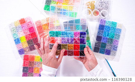 Flat lay. Woman hands gracefully poised over a collection of beads, sorted by color in transparent organizers. The array of beads spans a vibrant spectrum, from deep purples to bright oranges 116390128