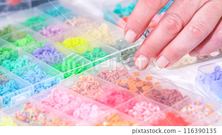 Woman hands gracefully poised over a collection of beads, sorted by color in transparent organizers. The array of beads spans a vibrant spectrum, from deep purples to bright oranges, meticulously 116390135