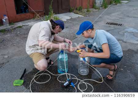 Dad and teenage son work together on the street near the garage. Outdoor activity. Family spend time. Helping and teach. Repairing a hose and pump for hand washing a car from a plastic bottle Dad and teenage son work together on the street near the garage. Outdoor activity. Family spend time. Helping and teach. Repairing a hose and pump for hand washing a car from a plastic bottle 116390659