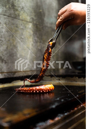 Hand of a cook catching an octopus tentacle with the tongs on a restaurant grill. Vertical 116393229