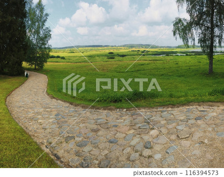 A serene outdoor landscape with a cobblestone path, bordered by grass and trees. Green fields, shrubs, and water in the distance under a partly cloudy sky.  116394573