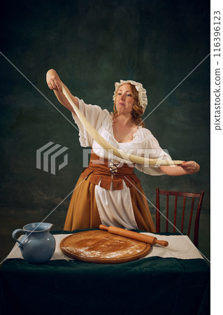 Woman in historical attire making dough for bread, set against dark, textured background. Medieval maid. Bakery, homemade bread 116396123